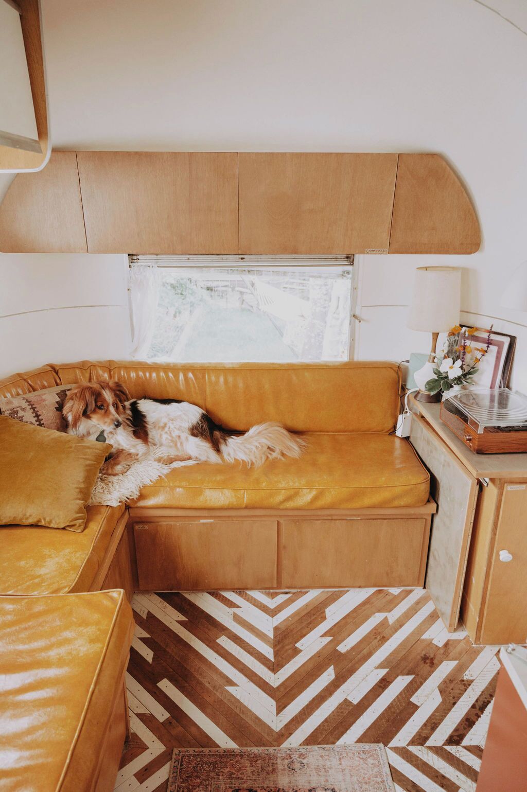 A cozy RV interior with a dog resting on a yellow couch, a wooden cabinet, and a patterned rug.