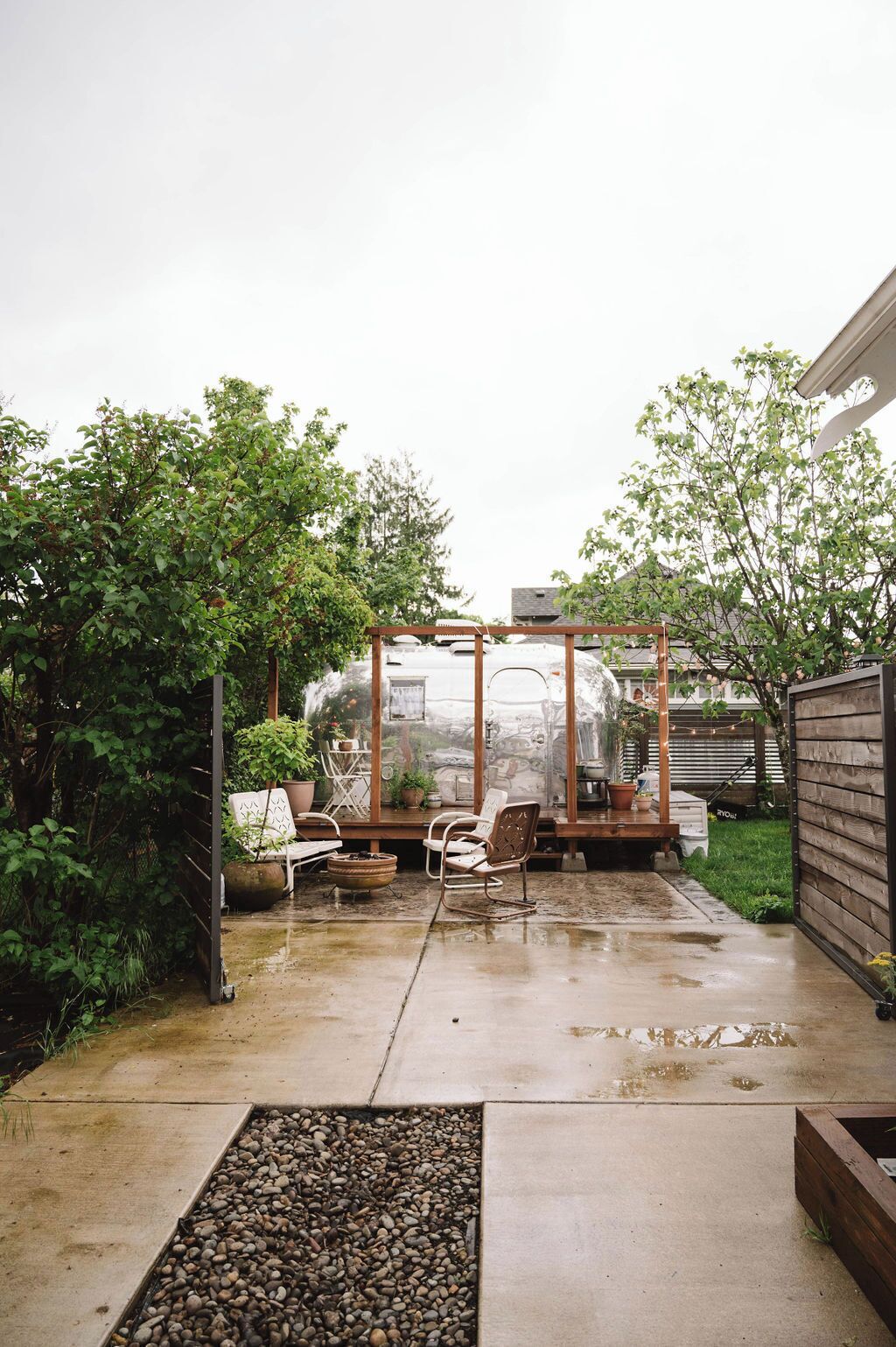 A covered patio area with a concrete floor, surrounded by greenery and a wooden fence.