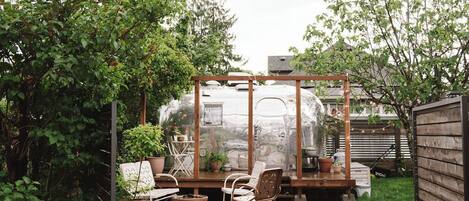A covered patio area with a concrete floor, surrounded by greenery and a wooden fence.