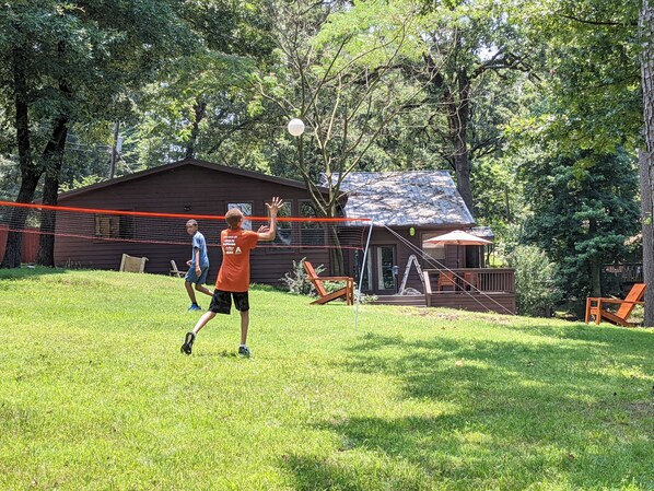 A grassy area with a volleyball net, a wooden cabin, and a person playing volleyball. - Sprawling 3-bedroom house in delightful Scroggins with WiFi, AC and lakeview (Scroggins)