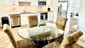 A modern kitchen with white cabinets, stainless steel appliances, and a glass-top dining table surrounded by patterned chairs.