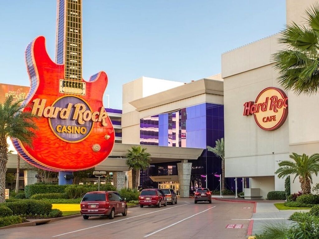 El edificio del casino Hard Rock con un llamativo letrero con forma de guitarra, una fachada moderna y un estacionamiento con automóviles.