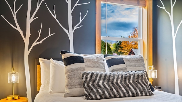 A neatly made bed with striped and textured pillows, a bedside lamp, and a view of autumn trees through a window. - Buck's Den - Snowmobile Trails, Trailer Parking, Near hiking, golfing, Sugarloaf (Eustis)