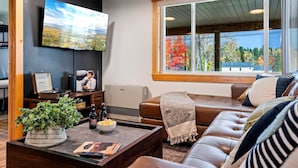 A modern living room with a large flat-screen TV, a comfortable leather sofa, a wooden coffee table, and a large window with a view of trees. - Buck's Den - Snowmobile Trails, Trailer Parking, Near hiking, golfing, Sugarloaf (Eustis)