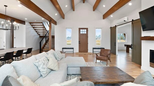 A spacious living area with a large sofa, a coffee table, and a fireplace. There is a staircase leading to an upper level and a dining area with chairs and a table.