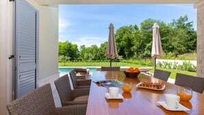 Una mesa de comedor con sillas de mimbre, una disposición para el desayuno con croissants y jugo de naranja, y vistas a una piscina y follaje.