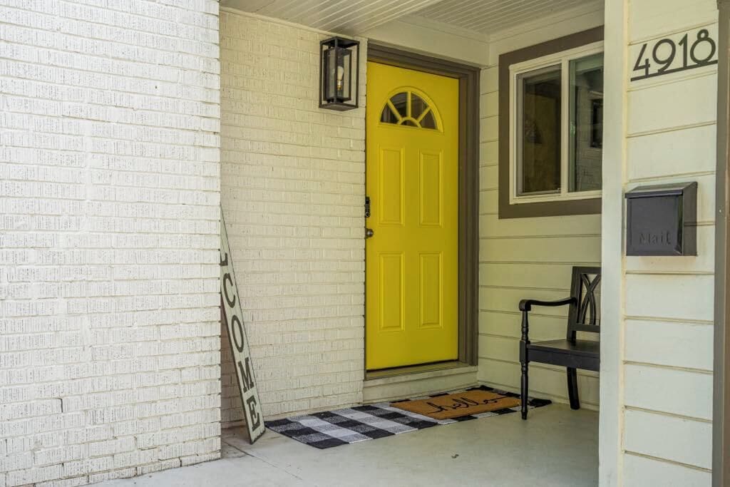 Une porte jaune surmontée d’une fenêtre, une boîte aux lettres et un banc devant une maison dont le mur est en briques blanches, et dont la boîte aux lettres porte le numéro 4918.