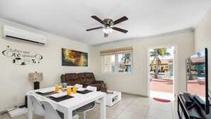 A modern living room with a dining area, a ceiling fan, an air conditioner, a TV, and a view of a pool area.