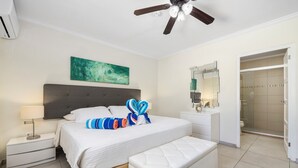 A hotel room with a bed, a bedside table, a nightstand, a bench, a ceiling fan, a bathroom with a shower, and a mirror.