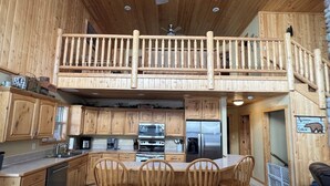 Una cocina de estilo rústico tipo cabaña, con armarios de madera, una isla central con sillas, un refrigerador de acero inoxidable y un balcón de madera con barandilla.