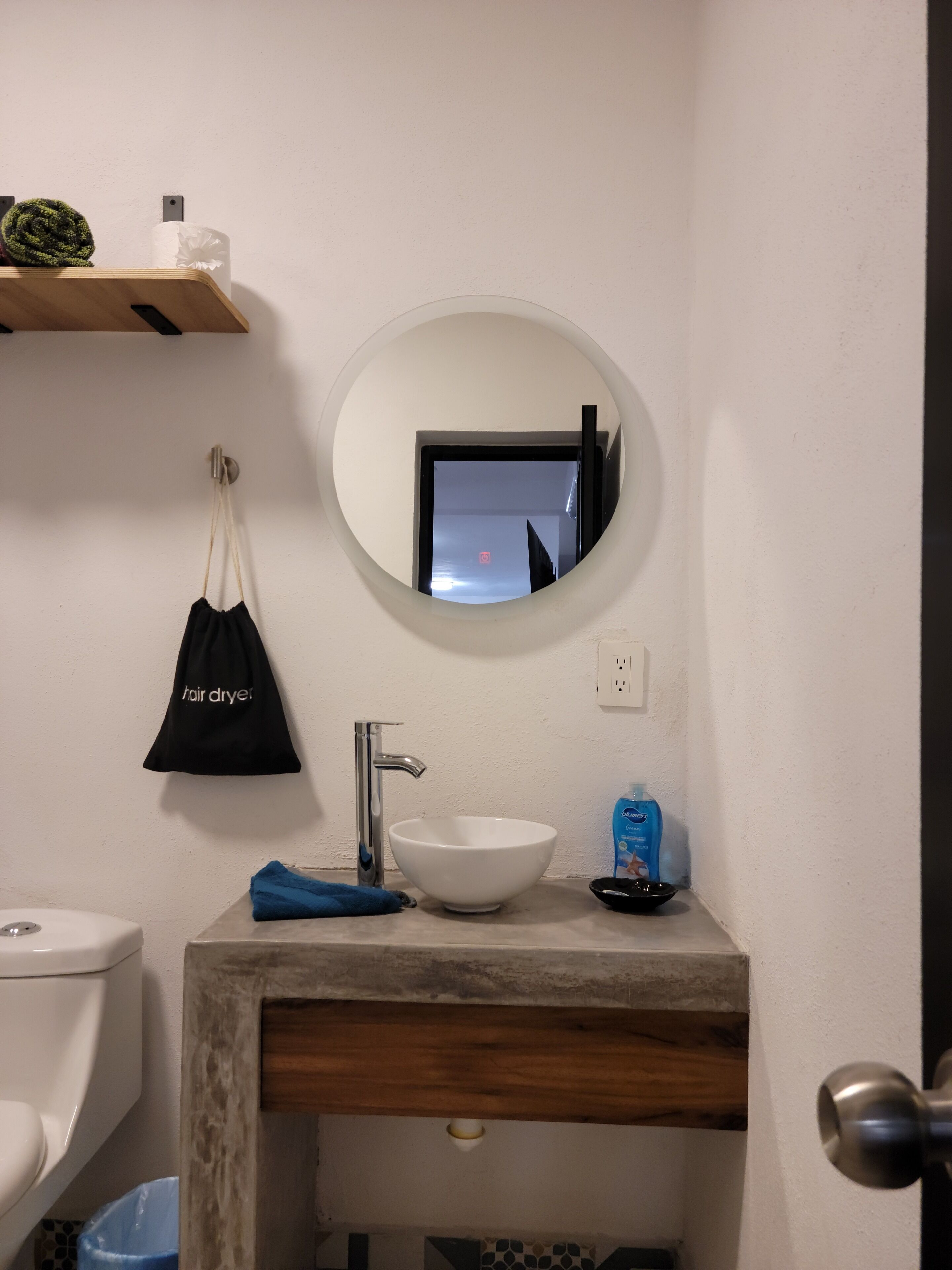 A bathroom with a white sink, a mirror, a hair dryer bag, and a blue trash bin.