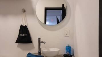 A bathroom with a white sink, a mirror, a hair dryer bag, and a blue trash bin.
