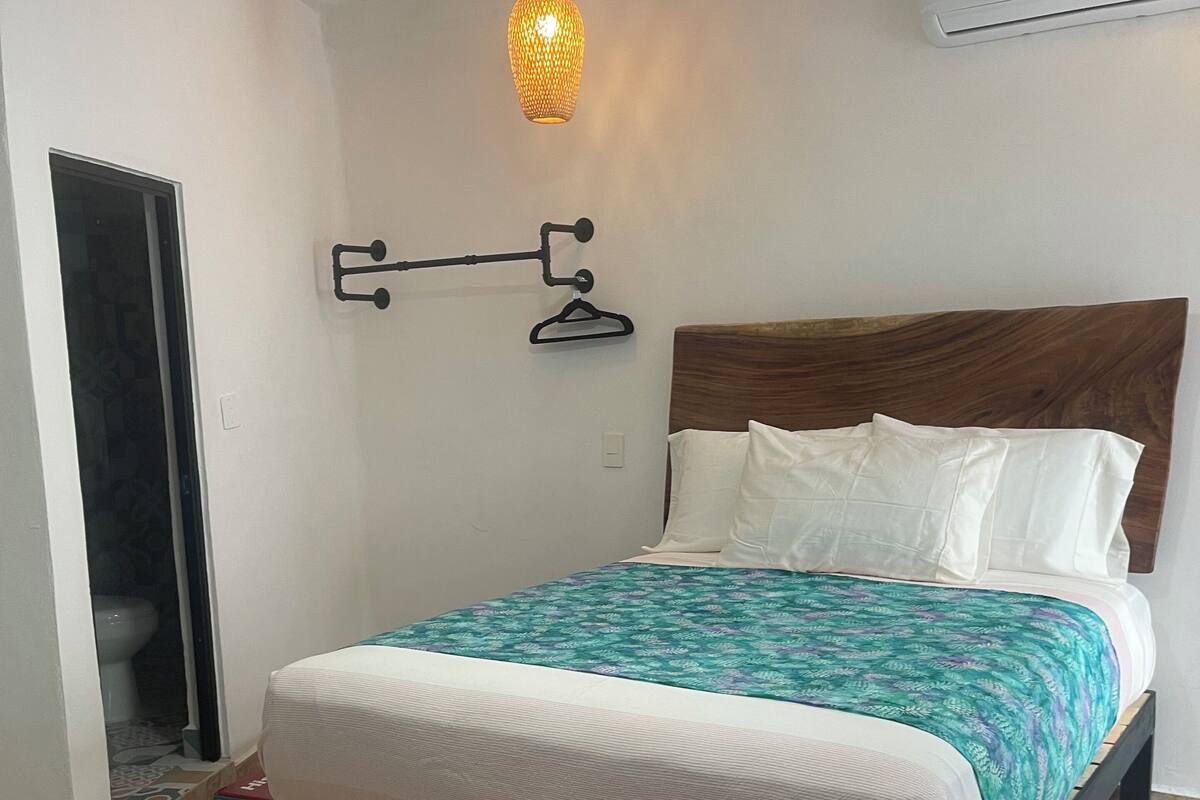 A bedroom with a bed, a hanging light fixture, a wall-mounted air conditioner, and a door leading to a bathroom.