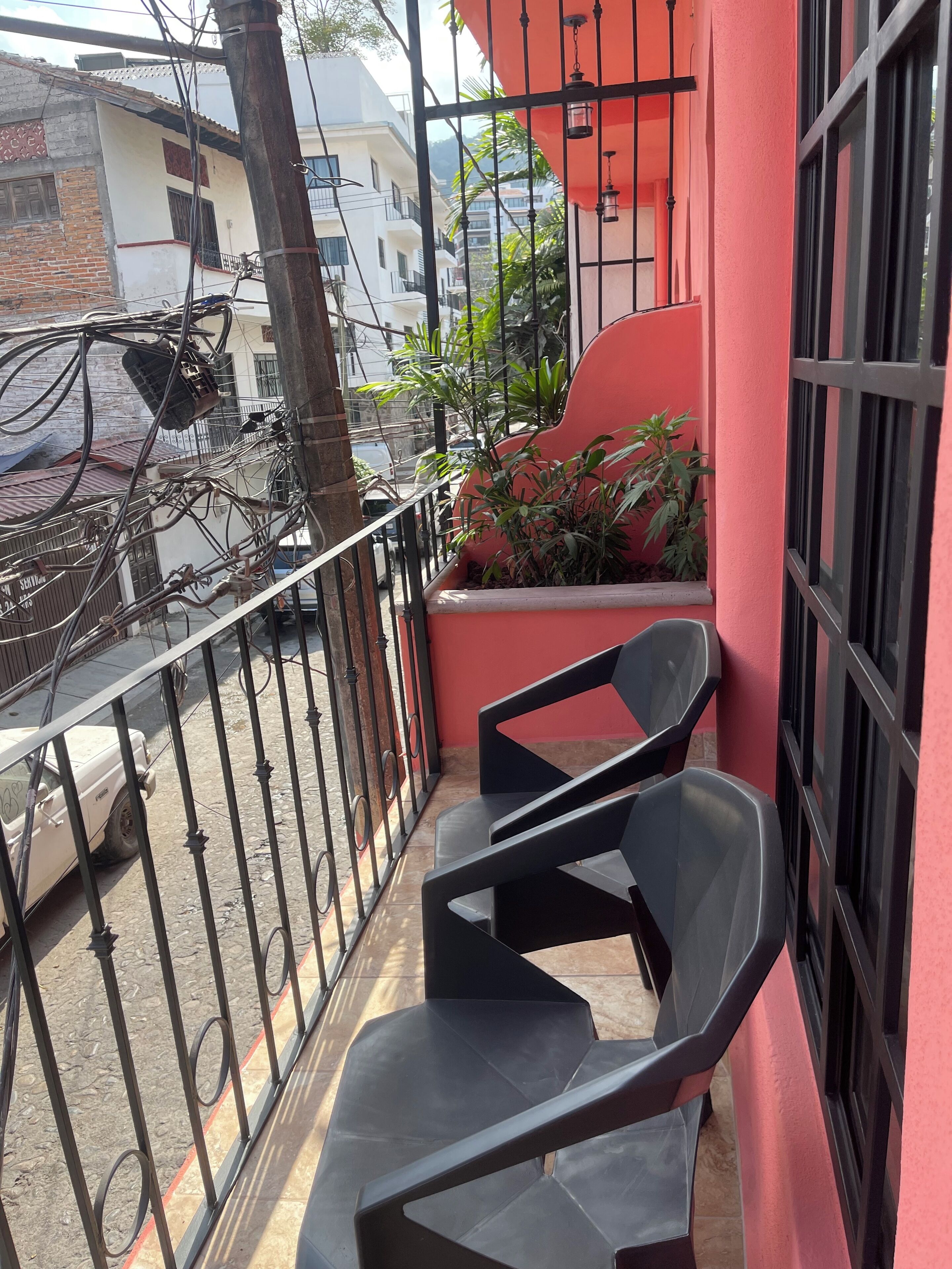 A balcony with a red wall, black metal railing, and two black plastic chairs. There are plants on the balcony and a white car parked outside.