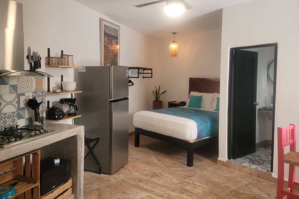 A compact kitchen with a refrigerator, stove, and microwave. A bed with a blue and white bedspread is in the adjacent room.