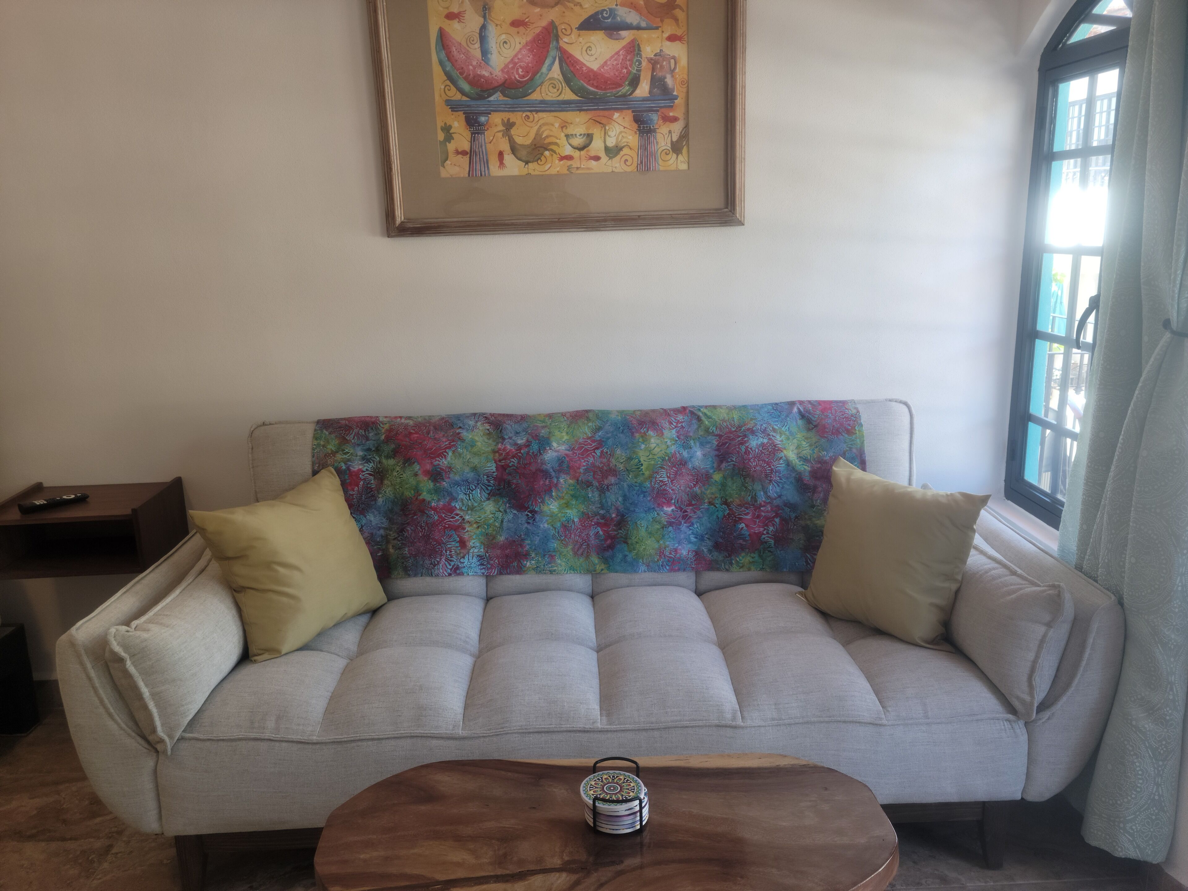 A living room with a sofa, a coffee table, a framed painting, and a window with curtains.