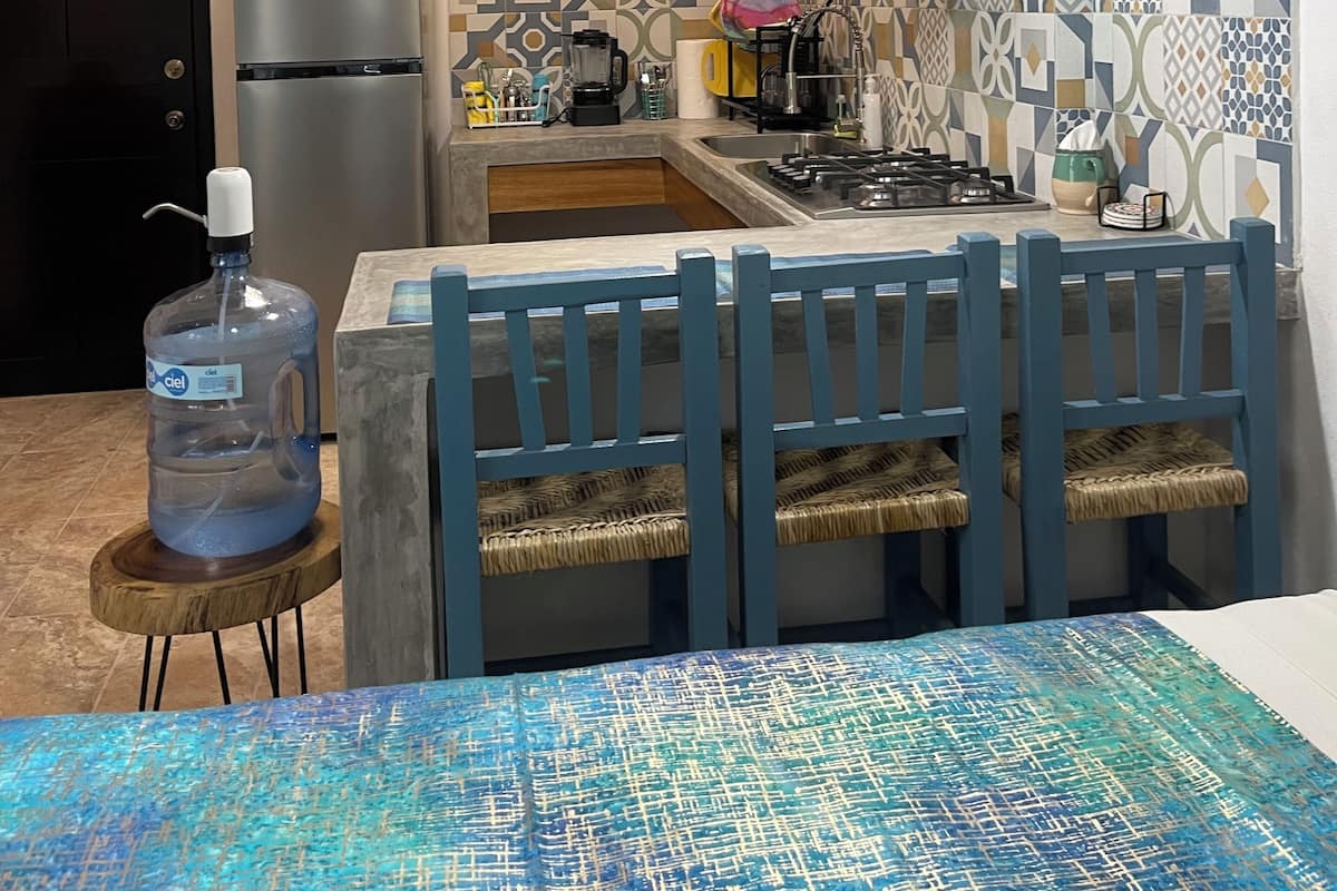A compact kitchen with a refrigerator, microwave, and gas stove. There is a large water dispenser and a dining area with a blue table and chairs.