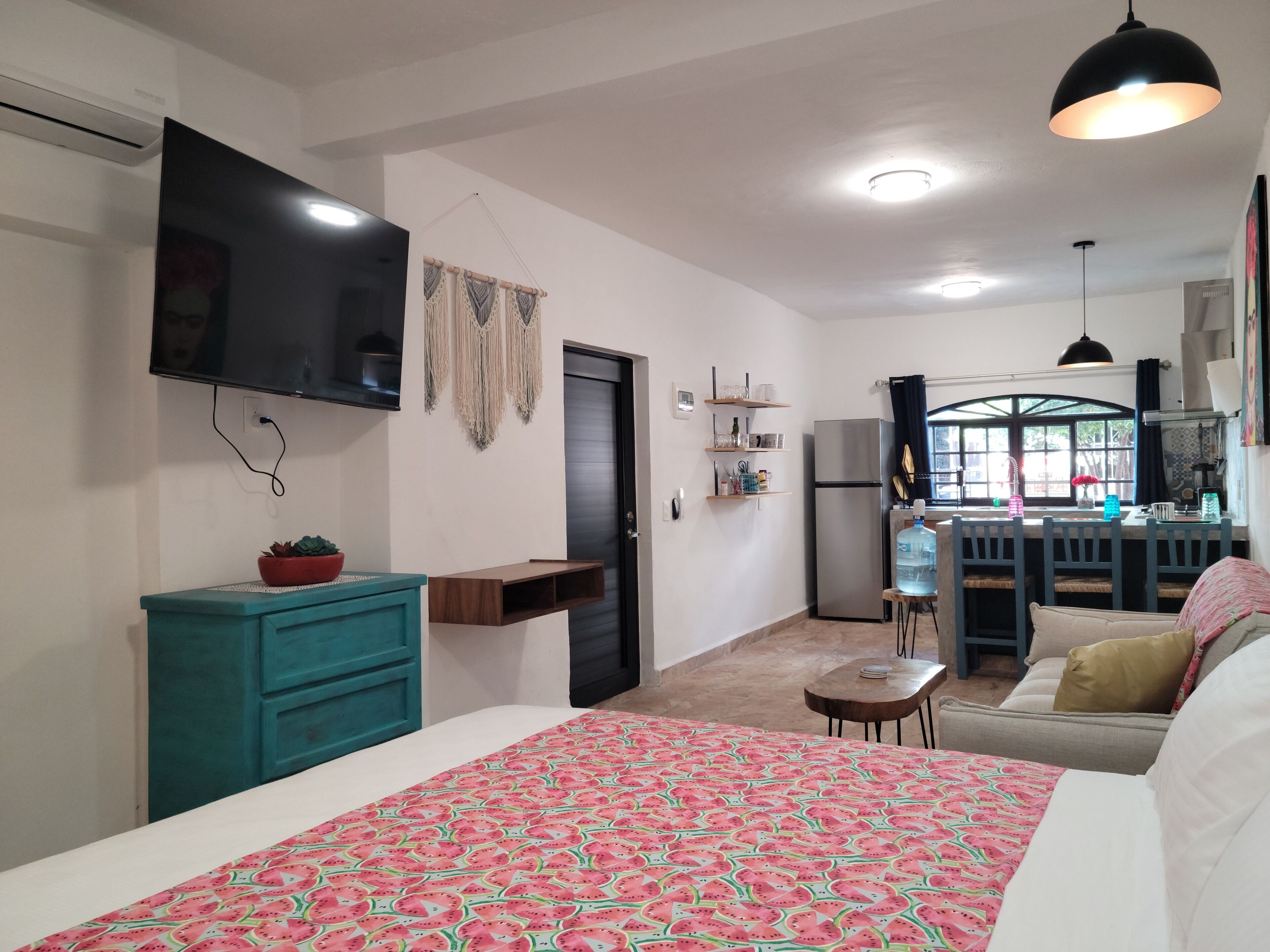 A compact living space with a bed, a TV mounted on the wall, a kitchenette, and a dining area with a table and chairs.