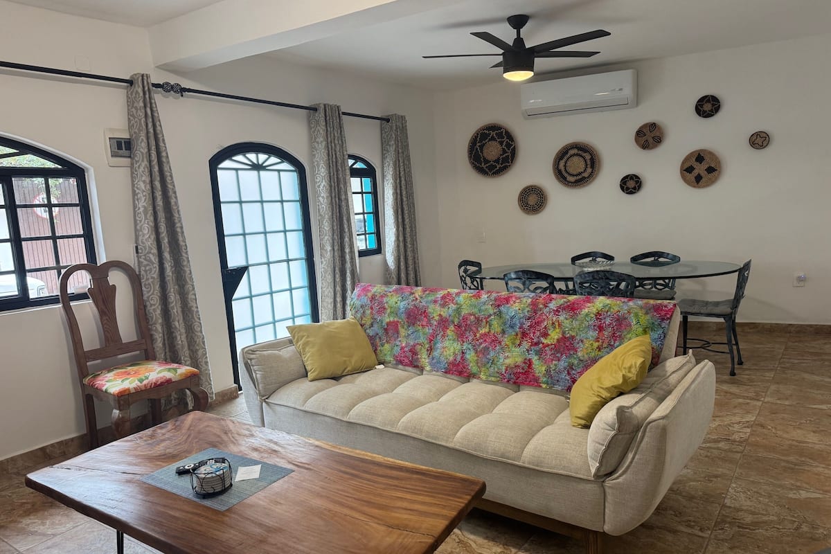 A living room with a sofa, a wooden coffee table, a dining table with chairs, and a ceiling fan.