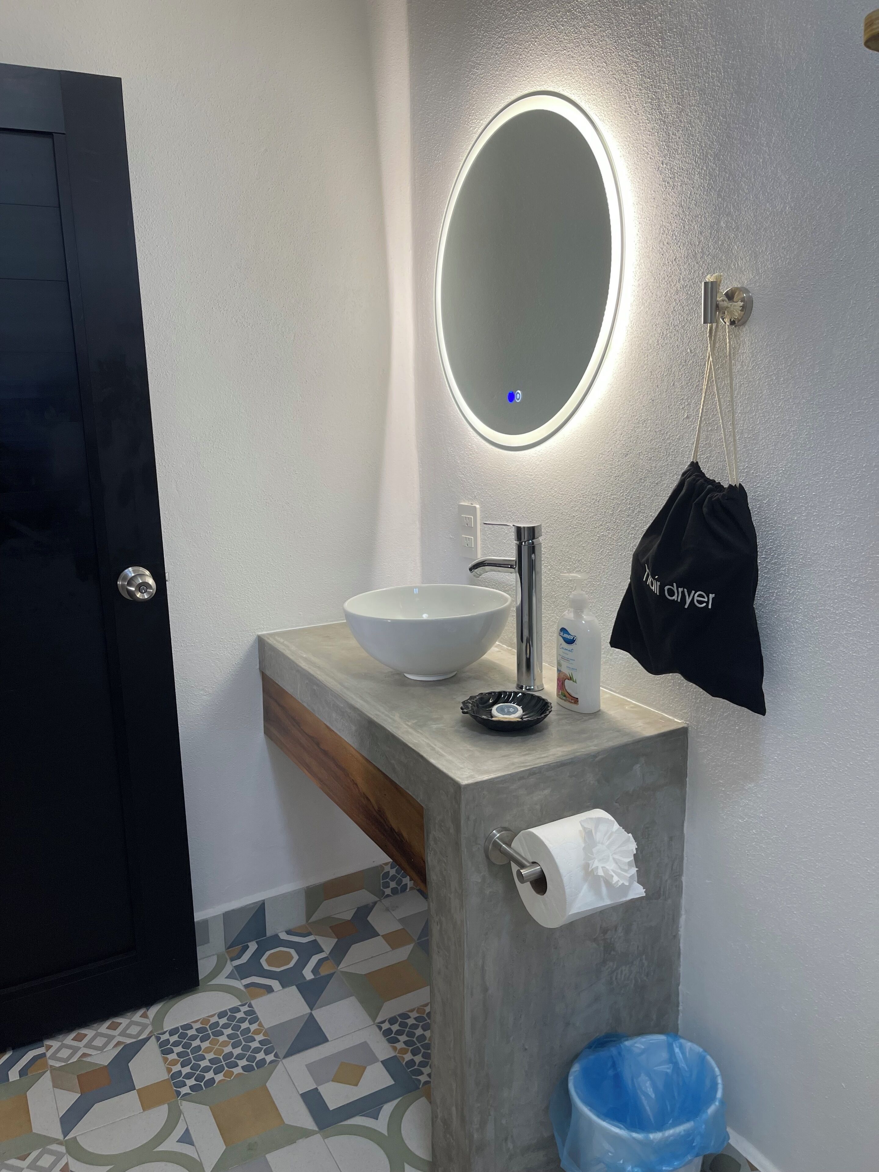 Een badkamer met een witte wastafel, een spiegel met verlichting, een handdoekrek en een blauwe prullenbak.