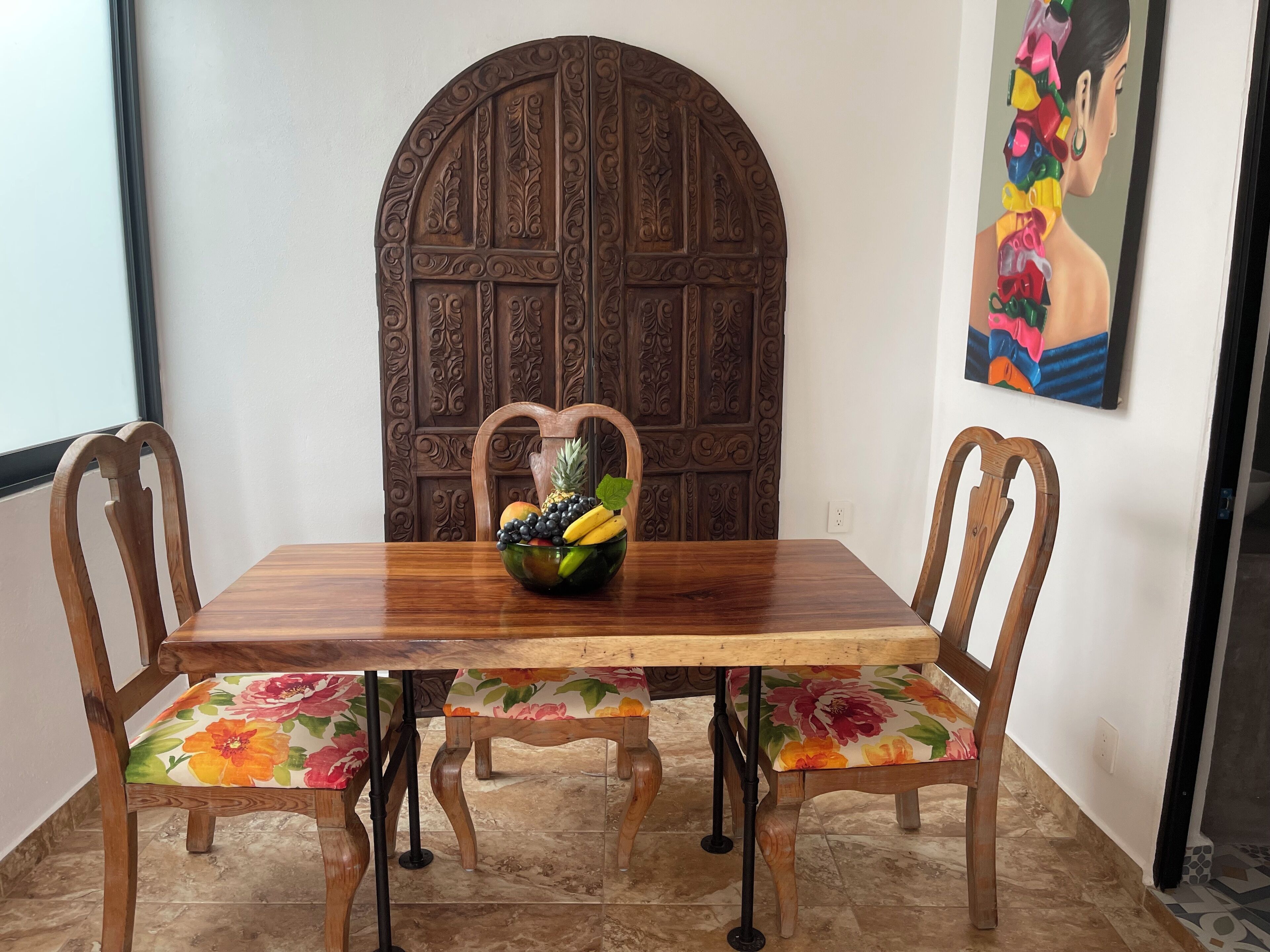 Una mesa de madera para comer con un frutero en el centro, rodeada de sillas de madera con tapicería floral.