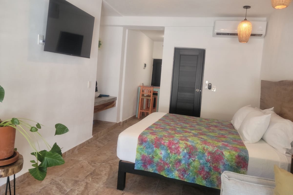 A bedroom with a bed, a TV mounted on the wall, a door, and a small table with a chair.