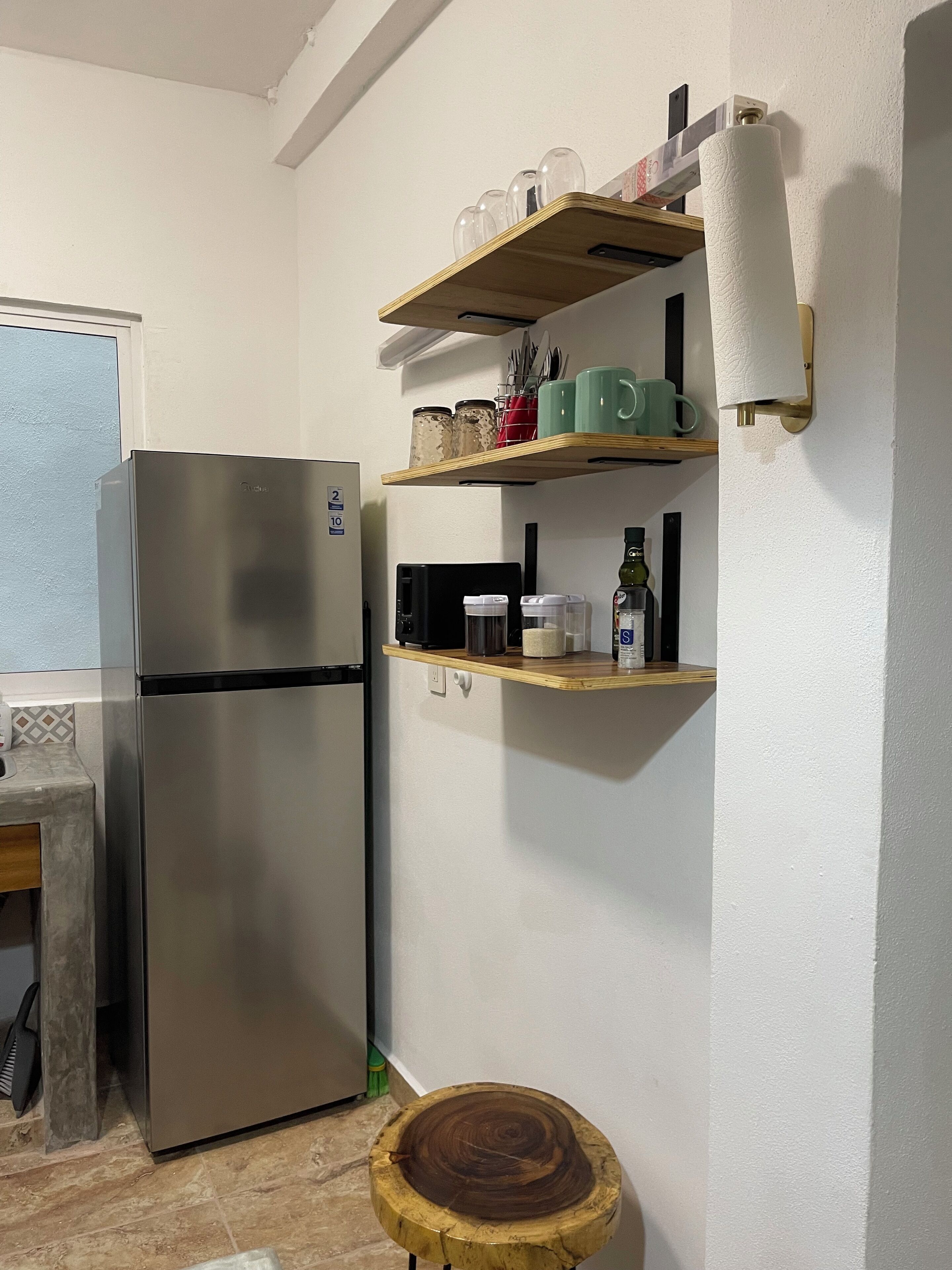 A kitchen with a stainless steel refrigerator, wooden shelves with kitchenware, and a round wooden stool.