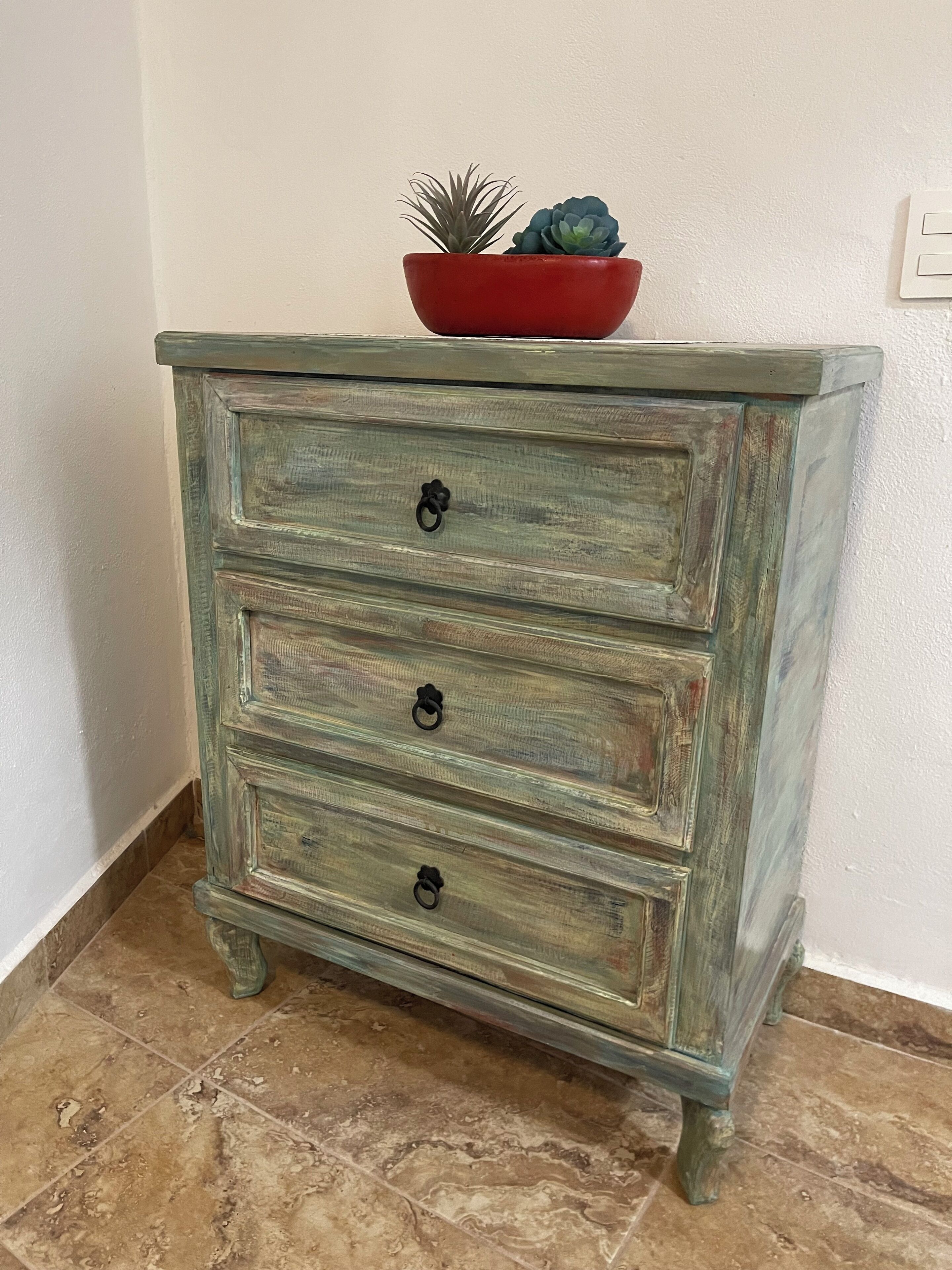 A rustic wooden chest of drawers with three drawers and a red bowl on top.