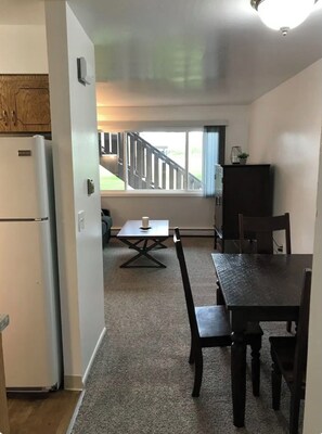 A compact kitchen with a refrigerator, a dining table with chairs, a small table with a candle, and a view of a wooden deck outside. - Ground Floor End Unit in Midtown Anchorage (Anchorage)