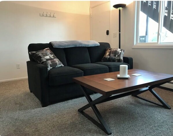 Un salon avec un canapé noir, deux coussins à motifs, une table basse en bois et un lampadaire.