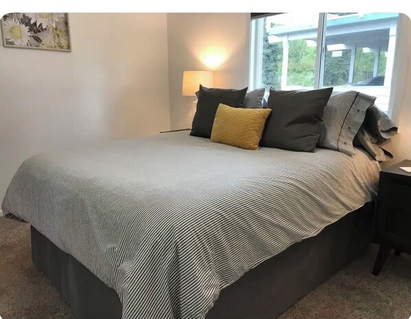 A neatly made bed with a striped bedspread and multiple pillows, a bedside lamp, and a window with a view of the outdoors. - Ground Floor End Unit in Midtown Anchorage (Anchorage)