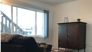 A living room with a dark-colored sofa, a wooden cabinet, and a window with a view of a staircase and parked cars. - Ground Floor End Unit in Midtown Anchorage (Anchorage)