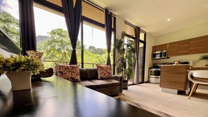 A modern living room with a dark wooden table, a brown sofa with patterned cushions, and a view of a kitchen and greenery outside. - Jaco Oasis Beach House (Jaco, Garabito)