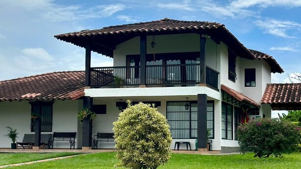 A two-story house with a balcony, a well-maintained lawn, and a small tree in the front yard. - Villa Privada eje Cafetero (Quindío)