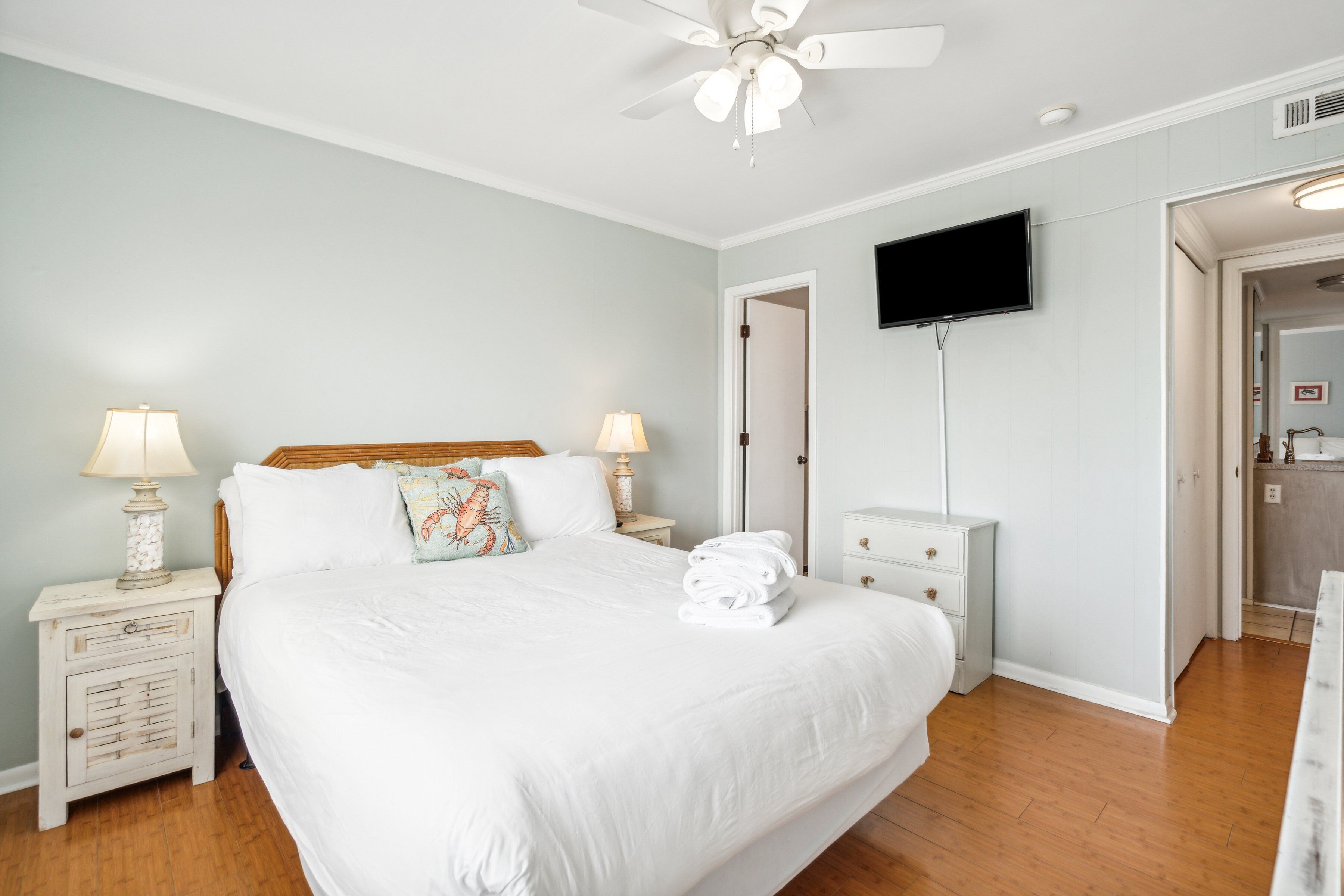 A bedroom with a large bed, two bedside tables with lamps, a ceiling fan, a TV mounted on the wall, and a bathroom visible through an open door.