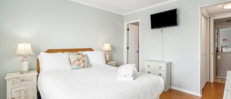 A bedroom with a large bed, two bedside tables with lamps, a ceiling fan, a TV mounted on the wall, and a bathroom visible through an open door.