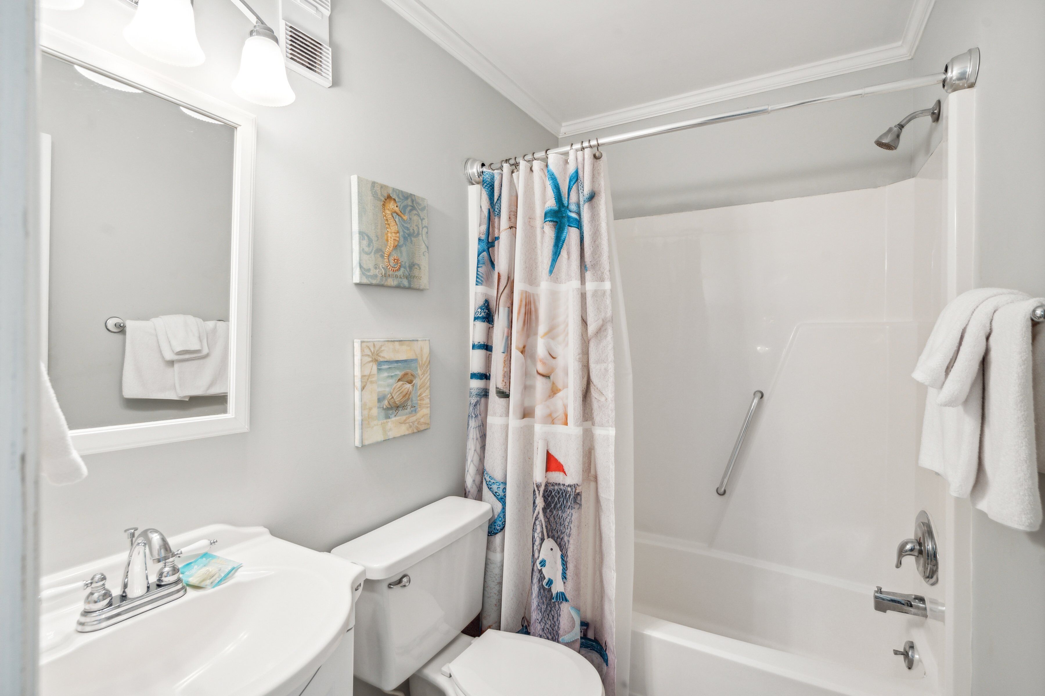 Un baño con lavamanos, inodoro y bañera blancos. Hay una cortina de ducha con motivos náuticos.