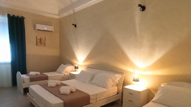 A hotel room with two beds, a bedside table, a lamp, an air conditioning unit, and a window with curtains.