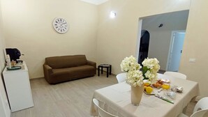 A dining area with a table set for two, a brown sofa, a small side table, and a clock on the wall.