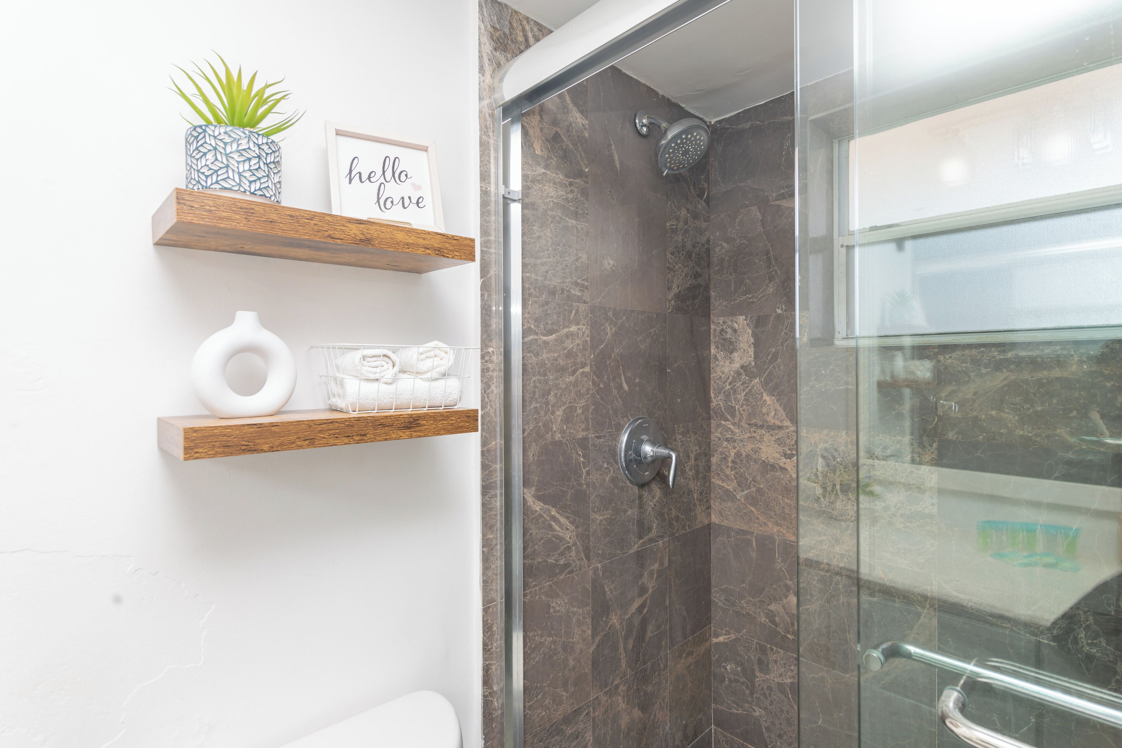 A modern bathroom with a glass shower enclosure, marble walls, and wooden shelves with toiletries.