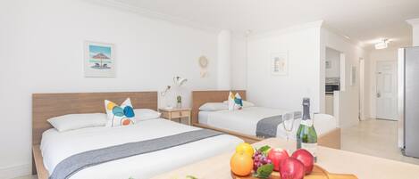 A hotel room with two beds, a dining table with fruit and a bottle, and a kitchen area visible in the background.