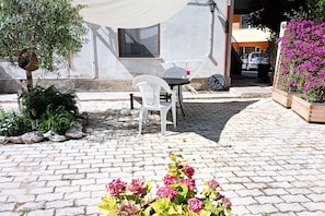Une terrasse avec une table et des chaises, une chaise blanche, une petite table et un pot de fleurs avec des fleurs roses.