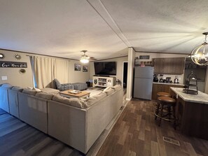 Un espacio habitable compacto con sofá, televisor, refrigerador y una zona de cocina con taburetes.