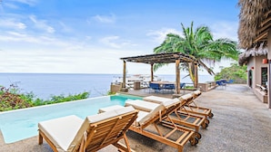 Zona de piscina con sillones de descanso, una pérgola y vista al océano.