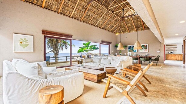 Amplia sala de estar con un gran ventanal con vista al mar, techo de paja y zona de comedor con muebles de madera.