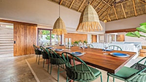 Comedor con una mesa de madera dispuesta para seis comensales, rodeada de sillas verdes, bajo dos lámparas colgantes tejidas.