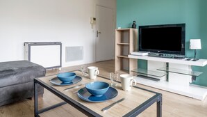 A modern living room with a sofa, a glass-top dining table with blue bowls and plates, a flat-screen TV on a white stand, and a wooden shelving unit. - Appartement Crista - Welkeys (Saint-Denis)