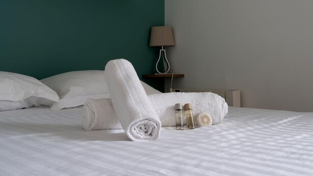 A neatly made bed with a rolled white towel, a bottle of lotion, and a round bath mat. - Appartement Crista - Welkeys (Saint-Denis)