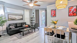 A modern living room with a black leather sofa, a dining table with chairs, a ceiling fan, and a large window with blinds.