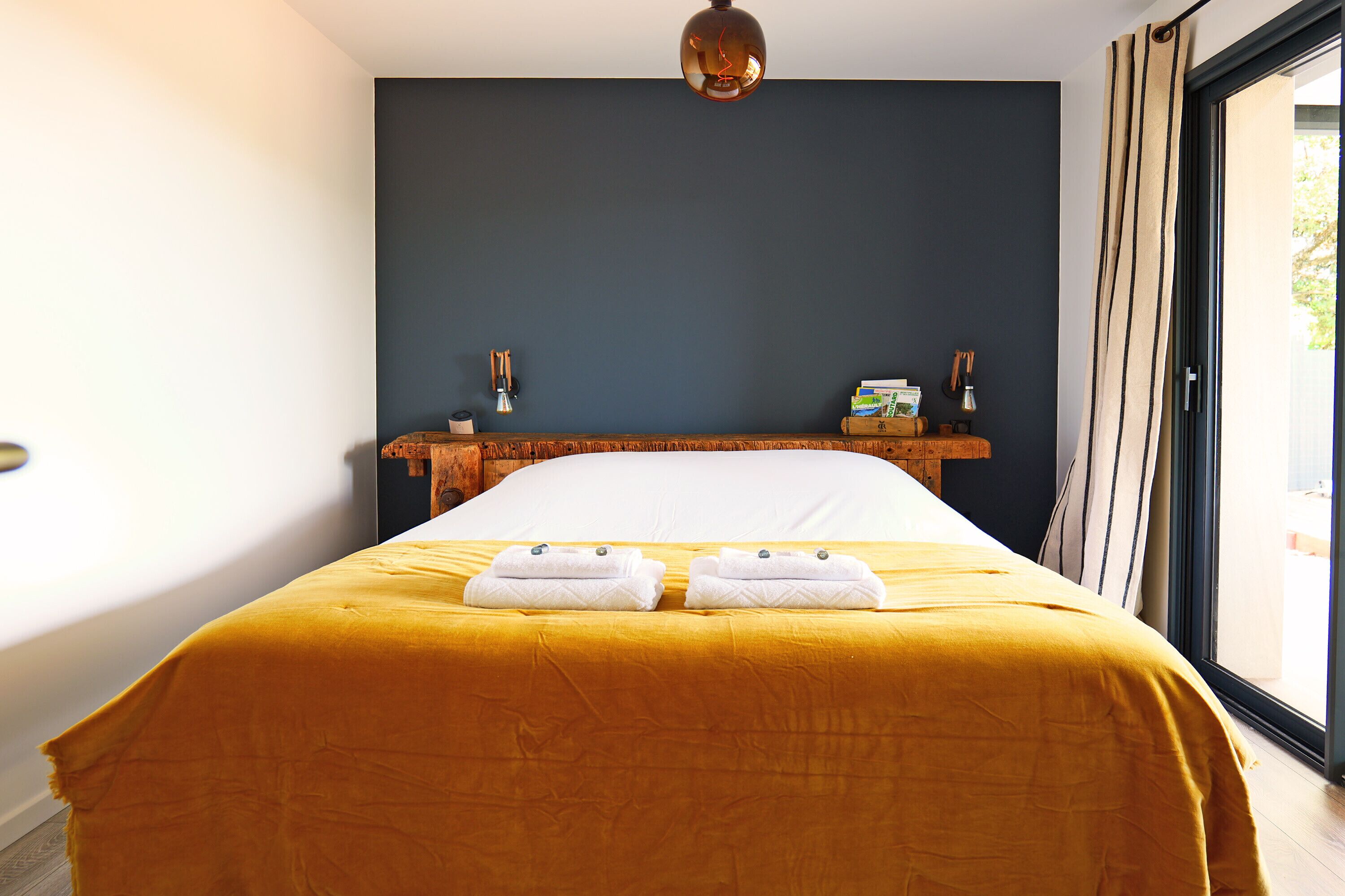 Une chambre avec un grand lit, une table de chevet en bois et une vue sur l’extérieur, visible à travers une porte vitrée.